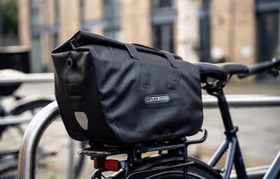 Black Trunk-Bag on a bike rack with a visible brand logo for Ortlieb attached to a bike, blurred urban background