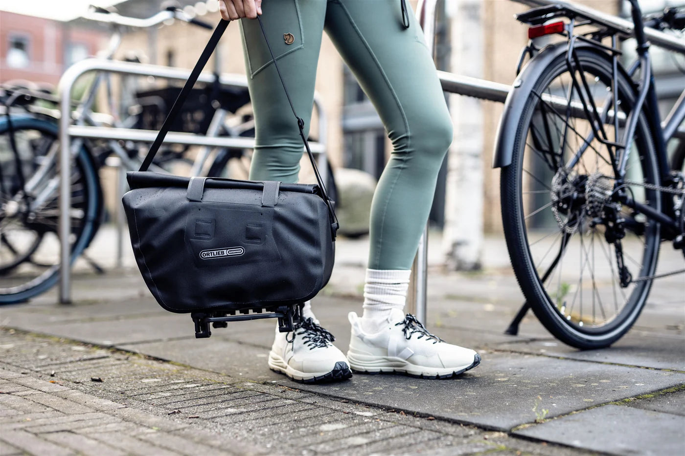 Person with black Ortlieb trunk-bag standing next to a bicycle on a sidewalk. Person holding trunk-bag by shoulder strap. 