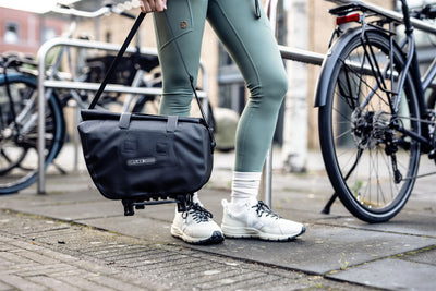 Person with black Ortlieb trunk-bag standing next to a bicycle on a sidewalk. Person holding trunk-bag by shoulder strap. 